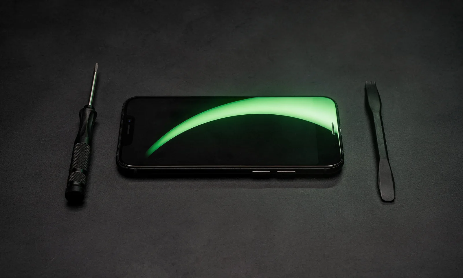 A phone on a repair workbench beside a precision screwdriver and a spudger, under a green diagnostic glow