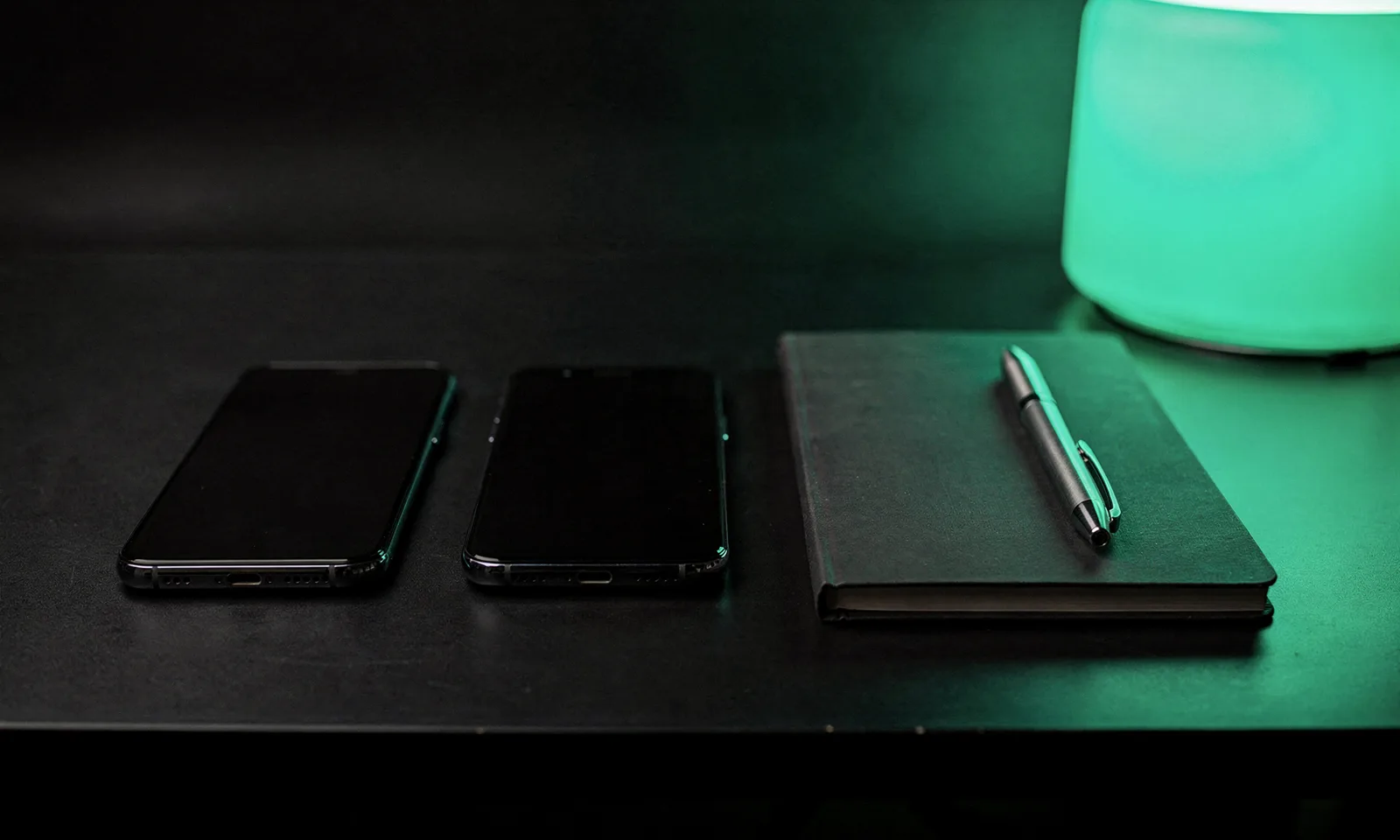 Phones lined up on a testing desk next to a notebook, lit by a green desk lamp